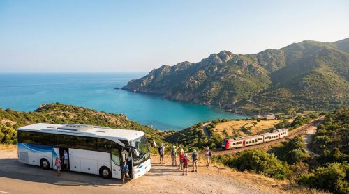 Reisegruppe an korsischer Küste steigt aus Bus aus. Im Hintergrund fährt ein Zug entlang einer Bucht und grünen Bergen.